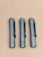 Three ReyLight Pineapple Mini Titanium MK3 flashlights with belt clips are lined up on a brown surface, ideal for everyday carry and equipped with high-CRI LEDs for outstanding illumination.