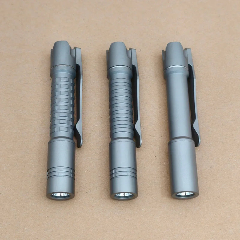 Three ReyLight Pineapple Mini Titanium - MK3 pen-style flashlights with pocket clips, designed for everyday carry, are arranged side by side on a plain brown surface.
