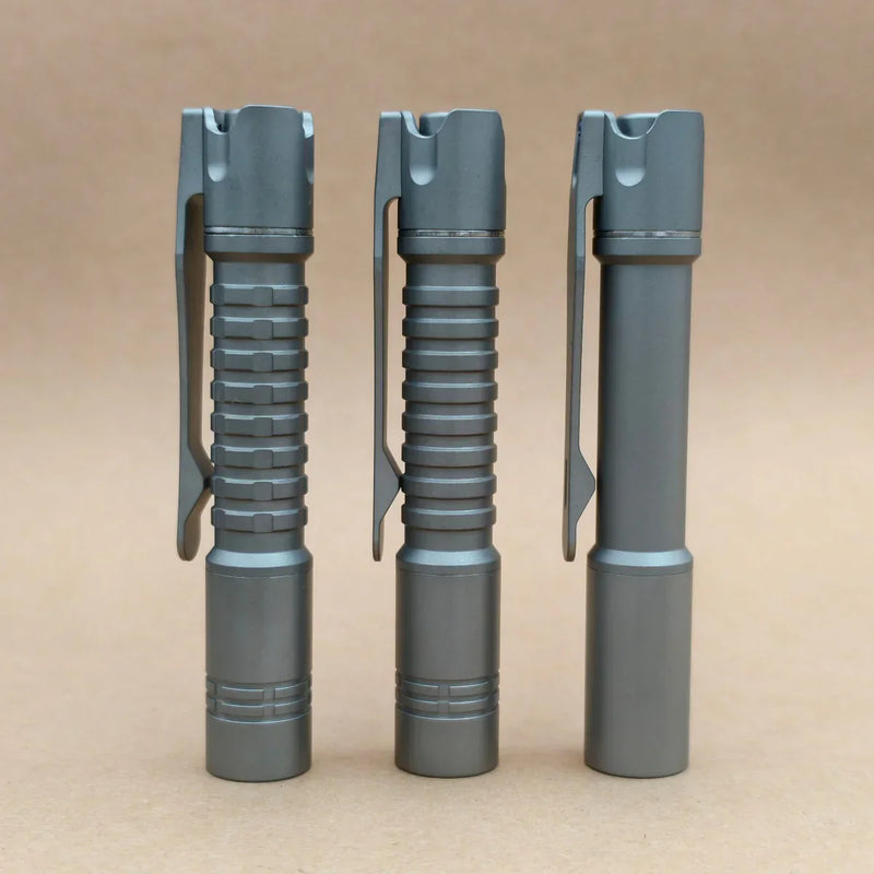 Three ReyLight Pineapple Mini Titanium - MK3 flashlights in gray, each with a pocket clip and unique grip texture, stand upright side by side on a plain light brown surface, showcasing their EDC-friendly design.