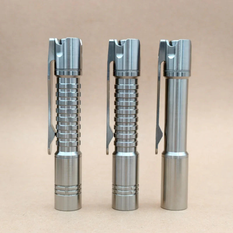 Three ReyLight Pineapple Mini Titanium MK3 flashlights with pocket clips stand upright on a beige surface, each featuring distinct grip textures and high-CRI LEDs—ideal for everyday carry.