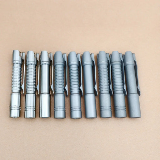 Nine metallic pens with varied textured grips line up on a light brown surface—perfect for your everyday carry, along with essentials like the ReyLight Pineapple Mini Titanium - MK3 flashlight.