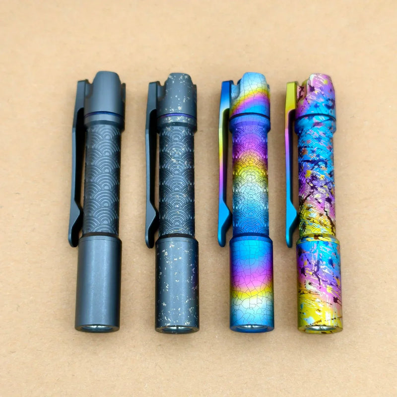 Four ReyLight Pineapple Mini Titanium Seigaiha - Anodized flashlights, each with distinct decorative patterns and colors, are lined up side by side on a plain brown surface.