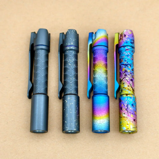 Four ReyLight Pineapple Mini Titanium Seigaiha - Anodized flashlights, each with distinct decorative patterns and colors, are lined up side by side on a plain brown surface.