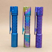 Three ReyLight Pineapple Mini Titanium Seigaiha - Anodized pens with pocket clips stand upright on a beige surface, each displaying a unique anodized finish and featuring the decorative Titanium Seigaiha engraved pattern.