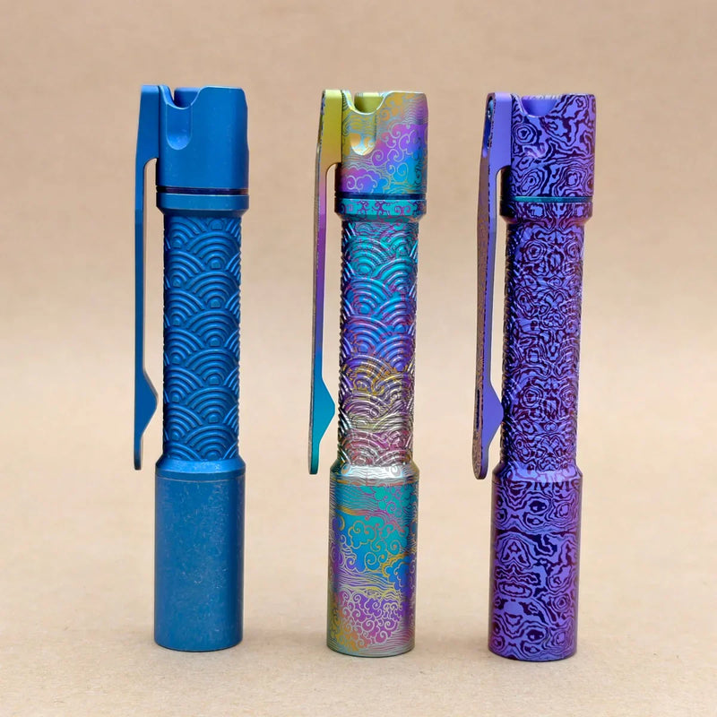 Three ReyLight Pineapple Mini Titanium Seigaiha - Anodized pens with pocket clips stand upright on a beige surface, each displaying a unique anodized finish and featuring the decorative Titanium Seigaiha engraved pattern.