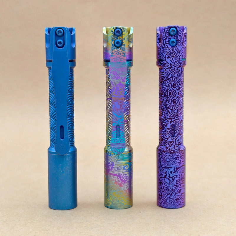 Three ReyLight Pineapple Mini Titanium Seigaiha - Anodized flashlights in blue, multicolor, and purple stand upright against a plain beige background, highlighting their unique Seigaiha-inspired titanium finishes.