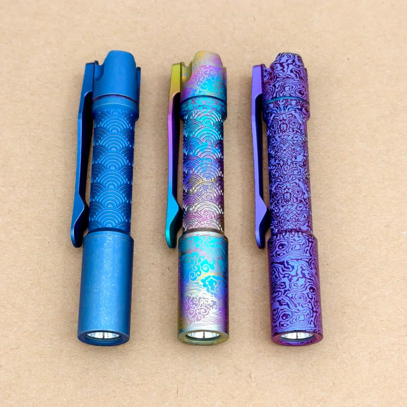 Three ReyLight Pineapple Mini Titanium Seigaiha - Anodized flashlights in blue, multicolored, and purple with intricate patterns and pocket clips are displayed side by side on a plain light brown surface.
