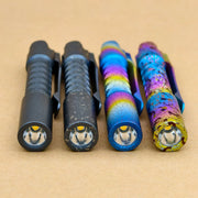 Four compact ReyLight Pineapple Mini flashlights—black, weathered black, anodized blue with rainbow accents, and the Titanium Seigaiha Anodized edition—are lined up on a brown surface.