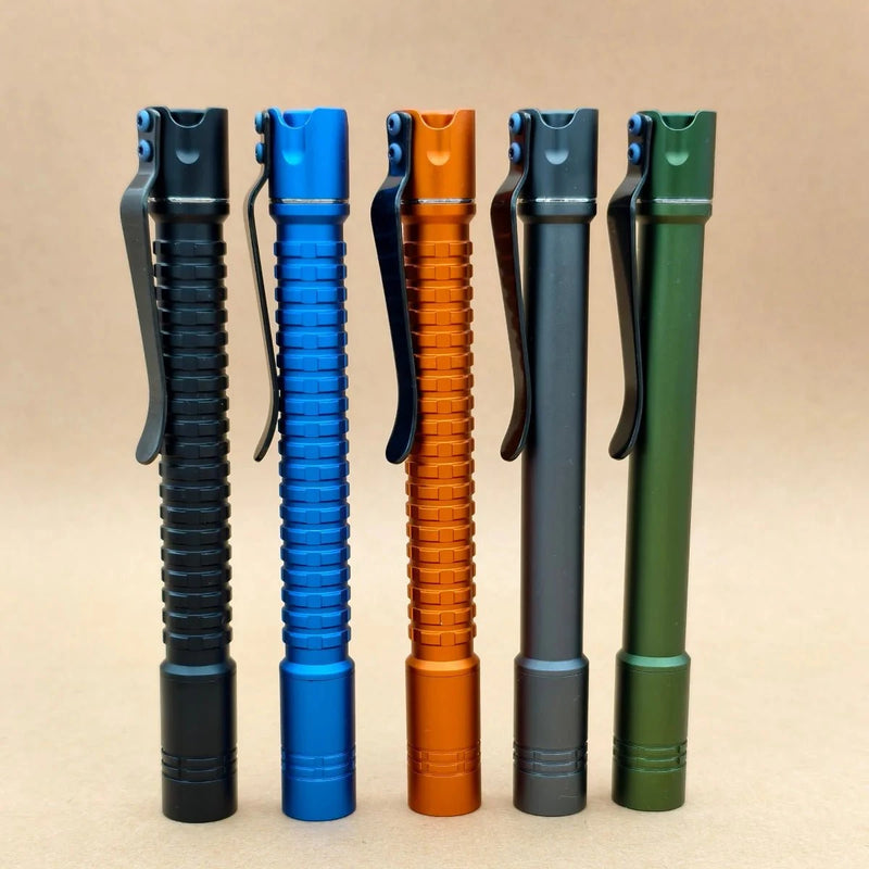 Five metal pen-shaped flashlights, including a rechargeable ReyLight Pineapple Penlight-MK II-Aluminum and a high-CRI LED flashlight, in black, blue, orange, gray, and green stand upright side by side on a light brown background.