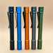 Five metal pen-shaped flashlights, including a rechargeable ReyLight Pineapple Penlight-MK II-Aluminum and a high-CRI LED flashlight, in black, blue, orange, gray, and green stand upright side by side on a light brown background.