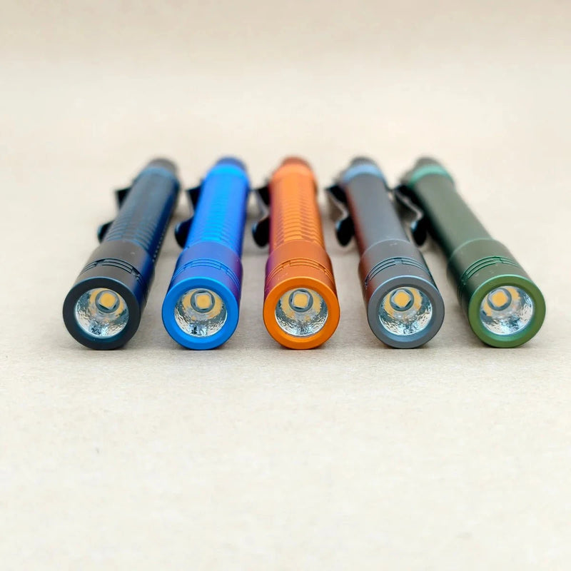 Five slim ReyLight Pineapple Penlight-MK II-Aluminum flashlights in black, blue, orange, gray, and green are arranged side by side on a beige surface, each high-CRI LED light shown facing forward.