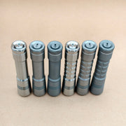 Six EDC flashlights, such as the ReyLight Pineapple Titanium, are lined up upright on a plain light brown surface, displaying different finishes and grip styles.