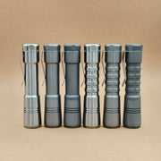 Six metallic EDC flashlights with pocket clips stand upright in a row against a plain tan background, including the ReyLight Pineapple Titanium for outstanding performance.