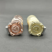 Two cylindrical objects with detailed caps, one copper and the other gold, resemble the ReyLight Rook - Brass flashlight's sleek design, placed side by side on a gray surface.