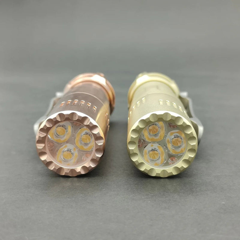 The Reylight Rook Brass flashlight, featuring three Nichia 519a LED bulbs, rests alongside another sleek metal-finished cylindrical flashlight on a gray surface.