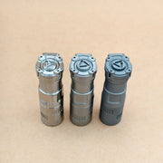 Three metal cylinders stand upright on a plain brown surface, each topped with a geometric symbol—these are ReyLight Rook - Ti flashlights, featuring a compact design, 18350 battery compatibility, and USB-C charging.