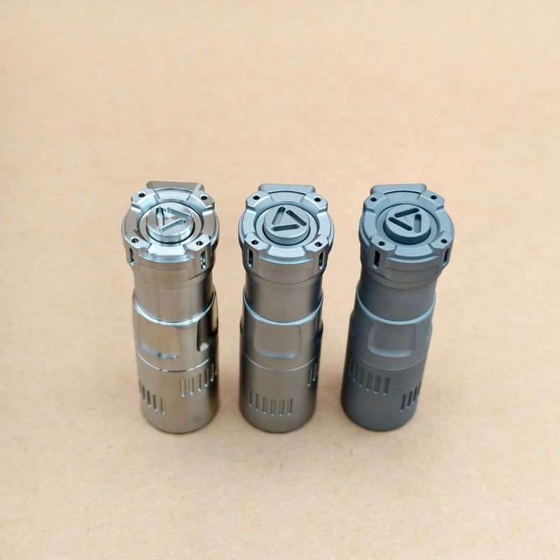 Three metal cylinders stand upright on a plain brown surface, each topped with a geometric symbol—these are ReyLight Rook - Ti flashlights, featuring a compact design, 18350 battery compatibility, and USB-C charging.