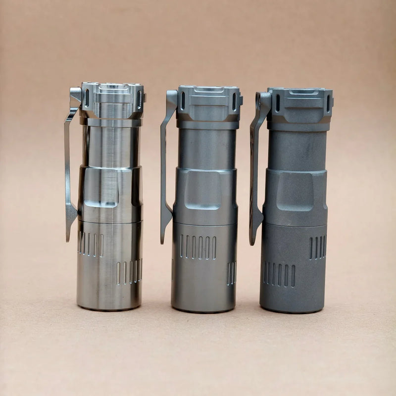 Three ReyLight Rook - Ti flashlights in polished, satin, and matte finishes stand upright in a row on a light brown background. Each compact flashlight runs on an 18350 battery for efficient performance.
