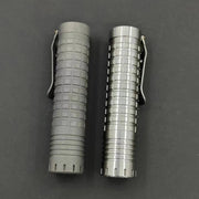 Two cylindrical metal objects with clips, featuring various textured patterns, lie adjacent on a dark surface. One features the distinctive charm of a ReyLight Ti Dawn Triple, highlighted by its Sapphire lens and Nichia 519A emitters.