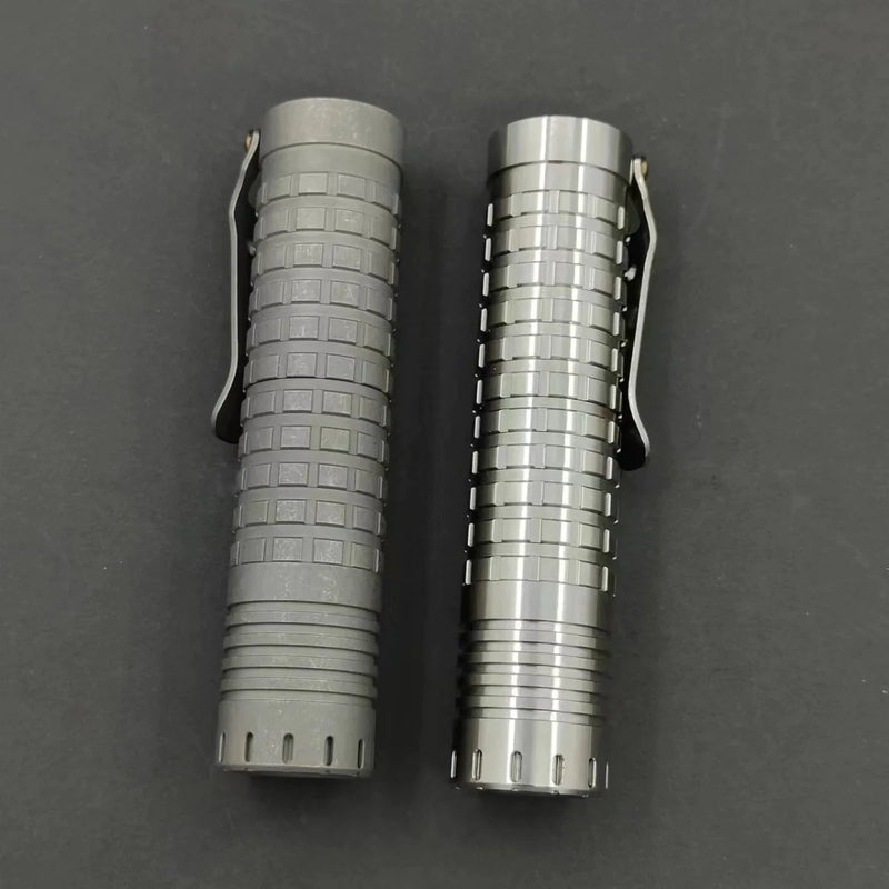 Two cylindrical metal objects with clips, featuring various textured patterns, lie adjacent on a dark surface. One features the distinctive charm of a ReyLight Ti Dawn Triple, highlighted by its Sapphire lens and Nichia 519A emitters.