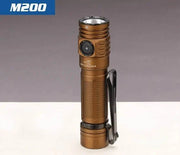 A bronze Skilhunt M200 V4 compact flashlight with a black side button, pocket clip, and magnetic charging stands upright on a white surface.