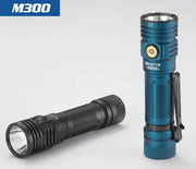 Two Skilhunt M300 V4 21700 flashlights, one upright in blue and one black lying on its side, are displayed on a plain light-colored surface.