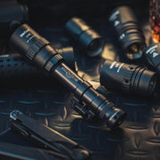 A close-up of multiple black tactical flashlights and tools, including the Weltool DF2E V2, displayed on a textured metal surface.