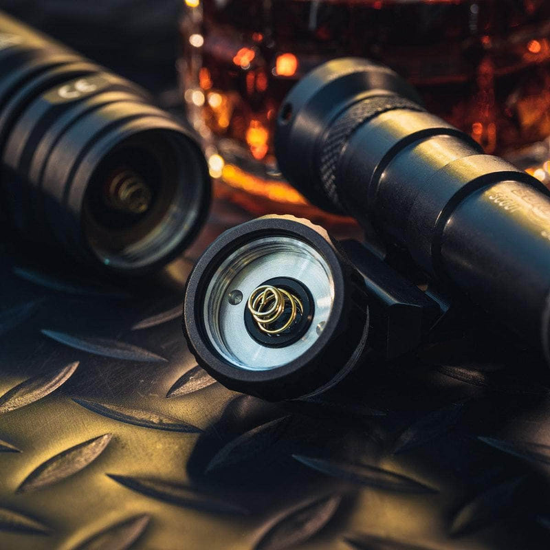Two disassembled black flashlights and a Weltool DF2E V2 adapter are shown on a textured metal surface, with a close-up of the battery compartment and a glass of amber liquid in the background.