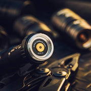 Close-up of the Weltool DF2E V2's tail end with an LH Series light head adapter nearby; other flashlights and a multitool are softly blurred in the background.