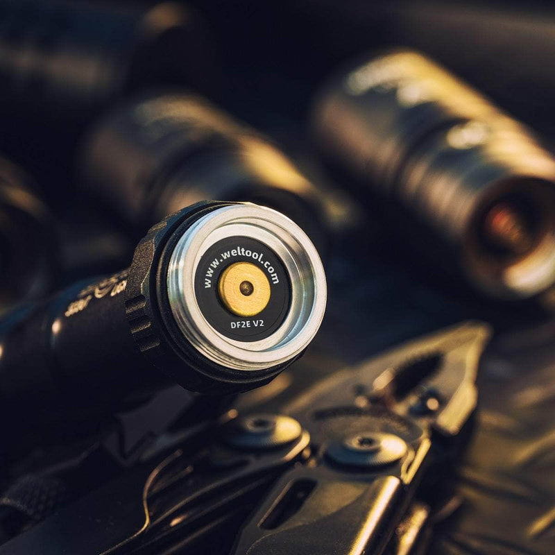 Close-up of the Weltool DF2E V2's tail end with an LH Series light head adapter nearby; other flashlights and a multitool are softly blurred in the background.