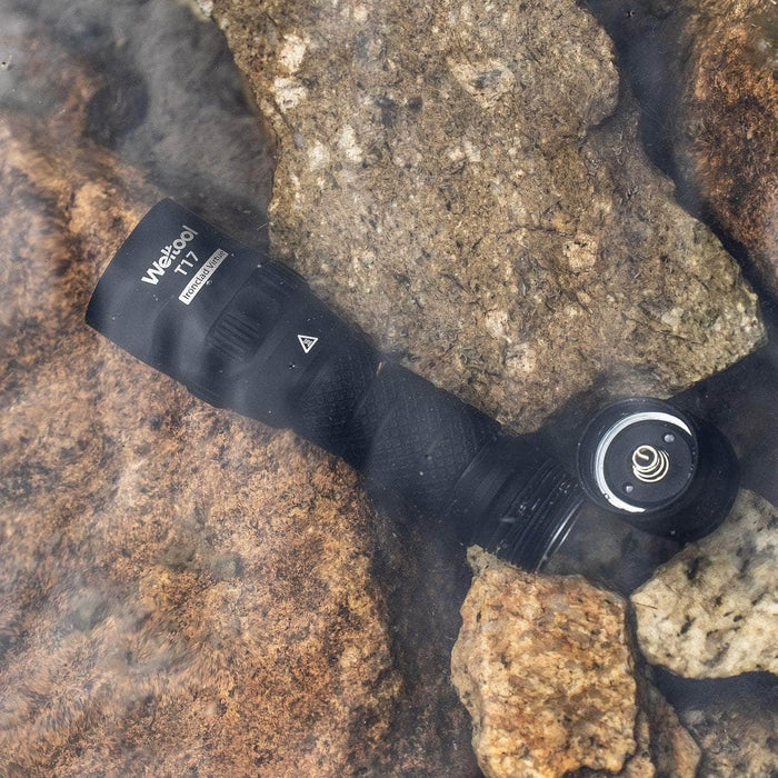 A durable Weltool T17 tactical flashlight laying on rocks in the water.