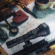 An assortment of outdoor and survival gear, including the Weltool F11R rechargeable long-range LED flashlight mounted on a firearm with a USB-C charging structure, a compass, a knife in a leather sheath, a map, and other tools are displayed on a wooden surface.