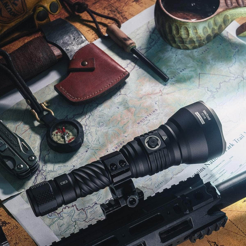 An assortment of outdoor and survival gear, including the Weltool F11R rechargeable long-range LED flashlight mounted on a firearm with a USB-C charging structure, a compass, a knife in a leather sheath, a map, and other tools are displayed on a wooden surface.