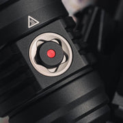 Close-up of the Weltool F11R, featuring a black, ridged component with a small red button in the center, surrounded by a metal ring. A triangular caution symbol is visible on the top left of the USB-C charging structure.