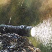 The Weltool F5, a high-lumen black LED flashlight, shines brightly even in heavy rain as it rests on a wet rock with water droplets glistening on its surface.