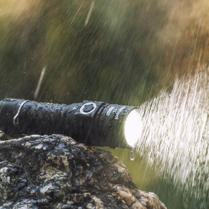 The Weltool F5, a high-lumen black LED flashlight, shines brightly even in heavy rain as it rests on a wet rock with water droplets glistening on its surface.