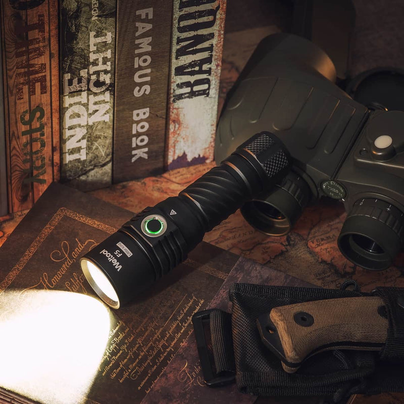 The Weltool F5, a high-lumen outdoor flashlight, illuminates a surface beside binoculars, a folding knife, and several books with vintage-style covers arranged in the background.