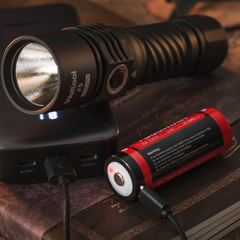 A Weltool F5 high-lumen flashlight in black and a red rechargeable battery are connected to a power bank via USB cables, resting on a wooden surface with books underneath.