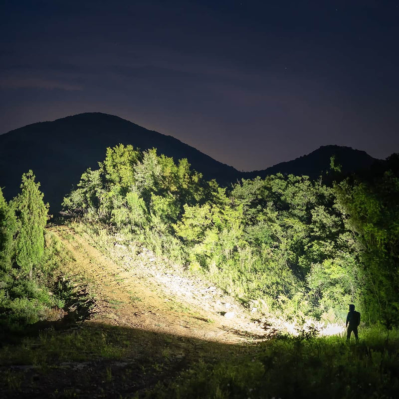 A person stands at night, illuminated by the powerful beam of the Weltool F5 flashlight, with dense trees and hills visible in the background beneath a dark sky.