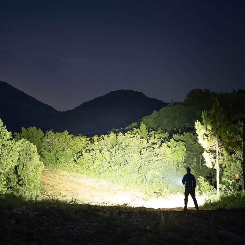 A person stands outdoors at night, illuminated by the Weltool F5 high-lumen LED flashlight, facing dense trees and hills under a dark sky.