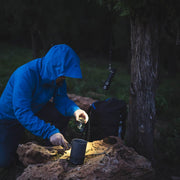 A person in a blue jacket prepares food on a rock outdoors at night, using a headlamp for light, while a Weltool F5 high-lumen LED flashlight hangs from a nearby tree.