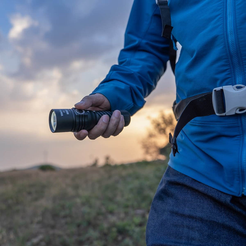 A person in a blue jacket holding the Weltool F5 high-lumen outdoor flashlight at sunset, with part of a backpack strap visible.