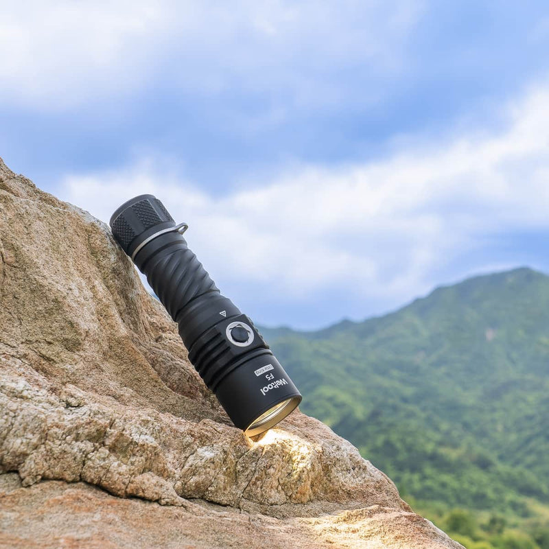 A Weltool F5 high-lumen black flashlight rests on a rocky surface with its beam shining, set against a backdrop of green hills and a cloudy blue sky.