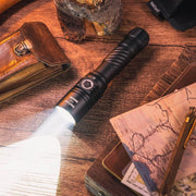 The Weltool F8R rechargeable LED flashlight casts a bright beam onto a wooden table with a wallet, map, notebooks, pen, and wood pieces—ideal for long-range lighting and quick USB-C charging.