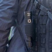 A black rechargeable LED flashlight labeled "Weltool F8R" is secured in a sheath attached to the strap of a black tactical backpack.