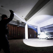 A person holding a Weltool F8R rechargeable long-range LED flashlight illuminates a white SUV and another person while walking in a covered parking area at night.
