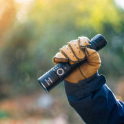 A person wearing a tan glove and dark sleeve holds a Weltool F8R rechargeable LED flashlight with USB-C charging outdoors, against a blurred natural background.