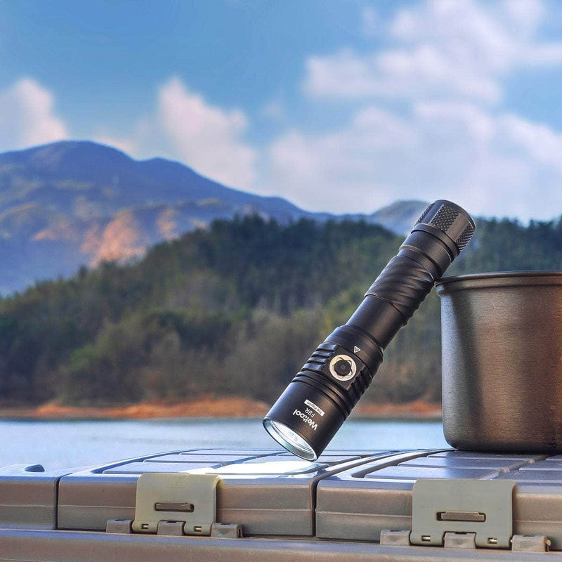 A Weltool F8R black rechargeable LED flashlight stands upright beside a metal pot on a plastic container outdoors, with a lake and mountains visible in the background.