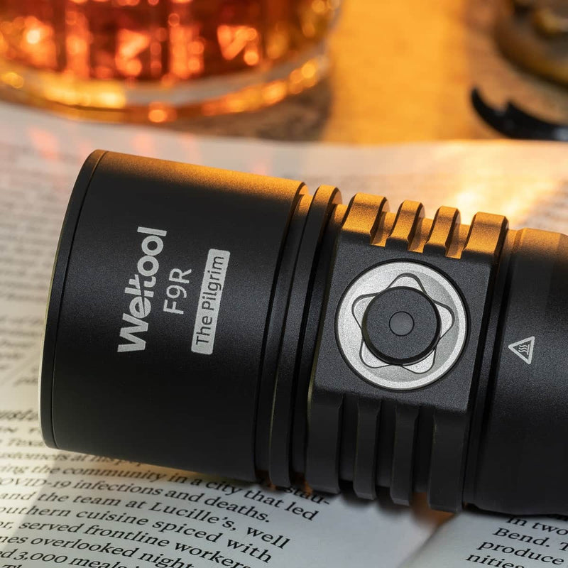 A black Weltool F9R high-lumen flashlight labeled "FOR The Pilgrim" rests on an open book, with a glass of liquid blurred in the background.