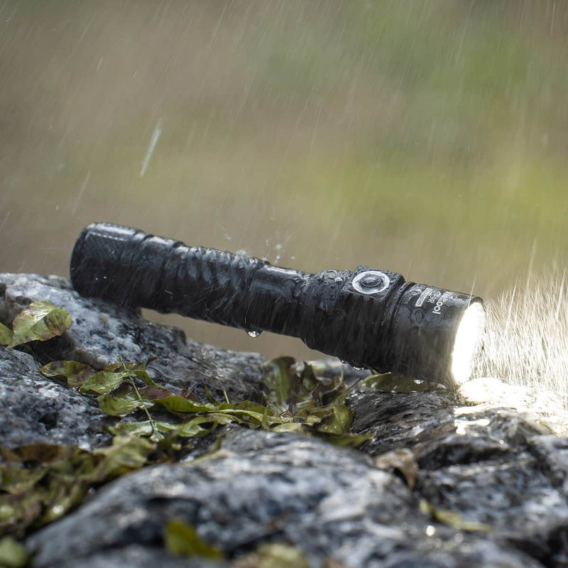 The Weltool F9R, a black rechargeable LED flashlight, is turned on and resting on wet rocks with rain falling and water droplets around it, surrounded by green leaves.